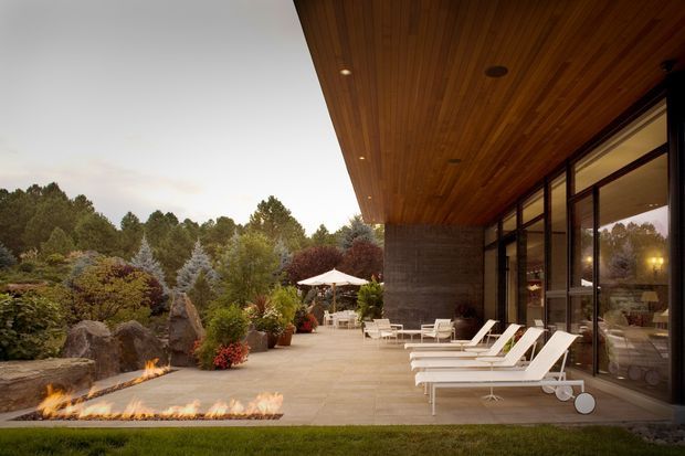 A sophisticated outdoor terrace beside a modern glass-walled building, featuring a broad timber-lined cantilevered roof, dark masonry wall, expansive glazed doors, pale stone paving, and a row of white sun loungers. A linear fire feature and landscaped garden with sculptural rocks, planting, and an umbrella seating area create a refined resort-like setting.