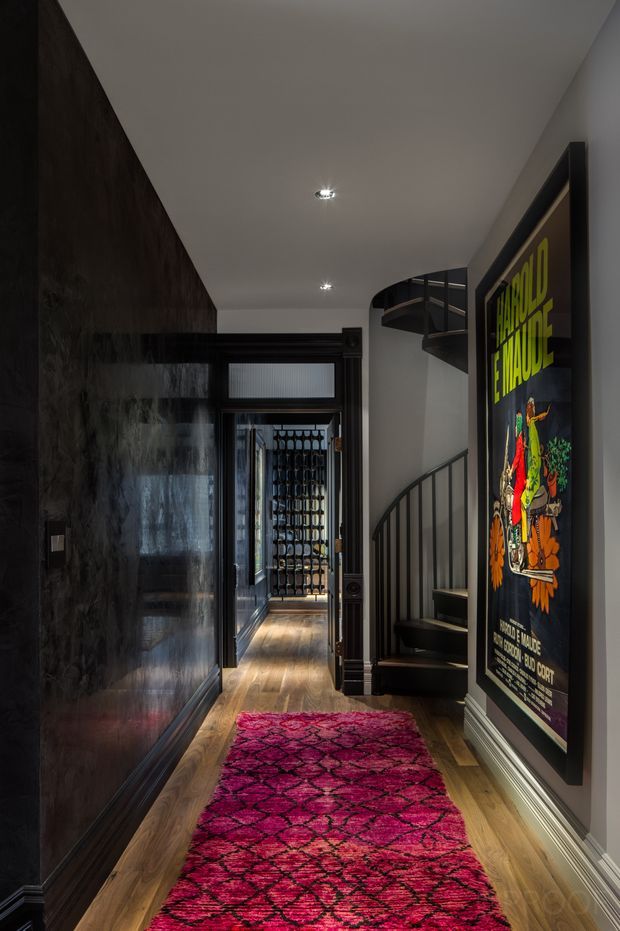 A narrow, dramatic residential hallway with warm timber flooring, dark glossy wall panelling on one side, white walls and ceiling, and recessed downlights. A vivid magenta patterned runner leads toward a glazed wine-storage area at the rear. On the right is a black-painted curved staircase with metal balustrading and a large framed vintage poster. The space has a moody, eclectic character with strong contrast between dark finishes and bright artwork and rug.