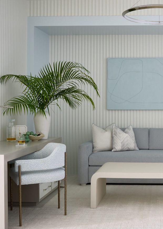 A softly lit contemporary living room with a pale grey sofa, sculptural cream coffee table, patterned upholstered armchair, and a light-toned console. Vertical fluted wall panelling creates a textured backdrop, complemented by a large abstract artwork, a potted palm, neutral cushions, and a circular modern pendant light. The overall palette is calm, cool, and refined.
