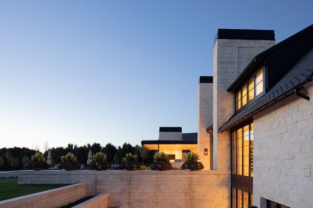 Exterior architectural photograph at dusk showing a contemporary multi-level residence composed of pale stone masonry walls, strong dark roof planes, tall chimney-like vertical elements, and large warm-glowing glazed openings. A landscaped terrace and low stone retaining wall occupy the foreground, with additional connected wings extending into the background.