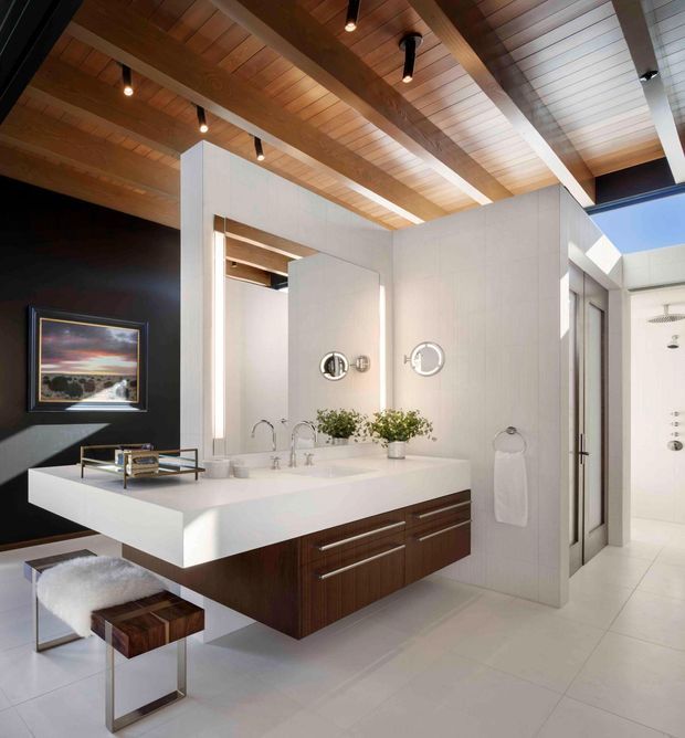 Contemporary bathroom with a floating double vanity, white integrated benchtop, dark timber drawers, large mirrors, and a frameless glass shower enclosure. The room features a dramatic black accent wall, white tiled surfaces, and a warm timber slatted ceiling with exposed beams and recessed spot lighting. A small upholstered stool and decorative plants add softness to the refined, high-contrast scheme.