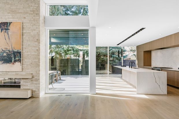 Bright open-plan contemporary kitchen and living area with floor-to-ceiling glazing opening to an outdoor terrace. The kitchen features a large white stone island, warm timber cabinetry, a white marble-look splashback, integrated linear lighting, and a sculptural black tap. The adjacent living area has pale timber flooring, a light textured brick feature wall, a slim fireplace, and minimal furnishings. Strong indoor-outdoor connection and abundant natural light.