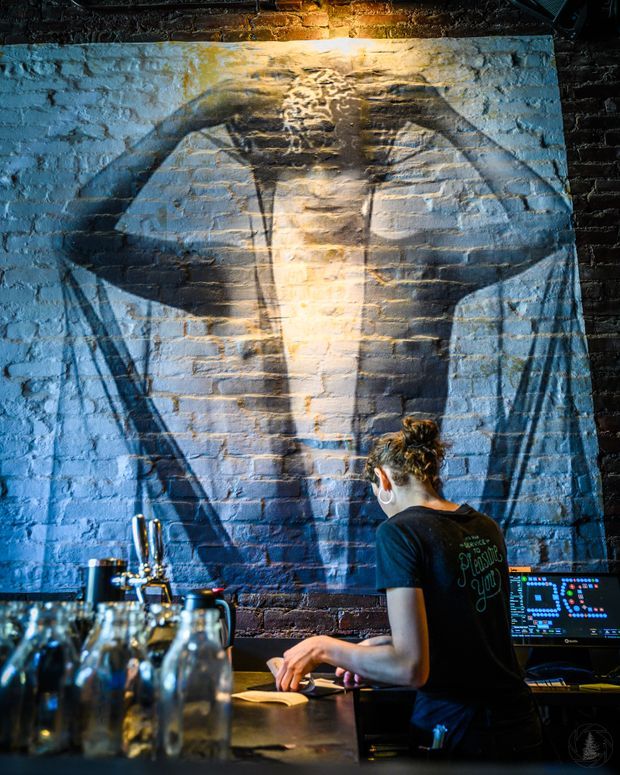 A dimly lit industrial bar interior with a bartender working behind a dark counter. The exposed brick wall is painted with a large blue-grey whale mural, while beer taps, glassware, bottles and a point-of-sale screen occupy the foreground. Warm pendant lighting contrasts with cool blue accent lighting, creating a dramatic hospitality atmosphere.