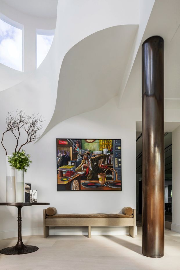 A bright, sculptural interior foyer or living space with sweeping curved white walls and ceiling forms, tall curved glazing, and a prominent cylindrical dark bronze or brown metal structural column. A minimalist upholstered bench sits beneath a colorful framed artwork, with a narrow dark console table and vase arrangement to the side. The pale floor and white surfaces create an airy, gallery-like atmosphere with strong natural light.