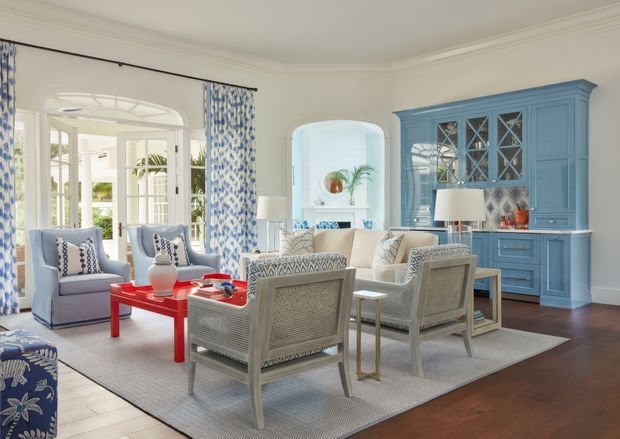 Bright, elegant living room with white walls and detailed crown moulding, dark timber flooring, blue built-in cabinetry, patterned blue-and-white curtains and upholstery, and a seating arrangement around a vivid red coffee table. Large glazed French doors and an arched opening bring in abundant natural light, creating a refined coastal-traditional atmosphere.