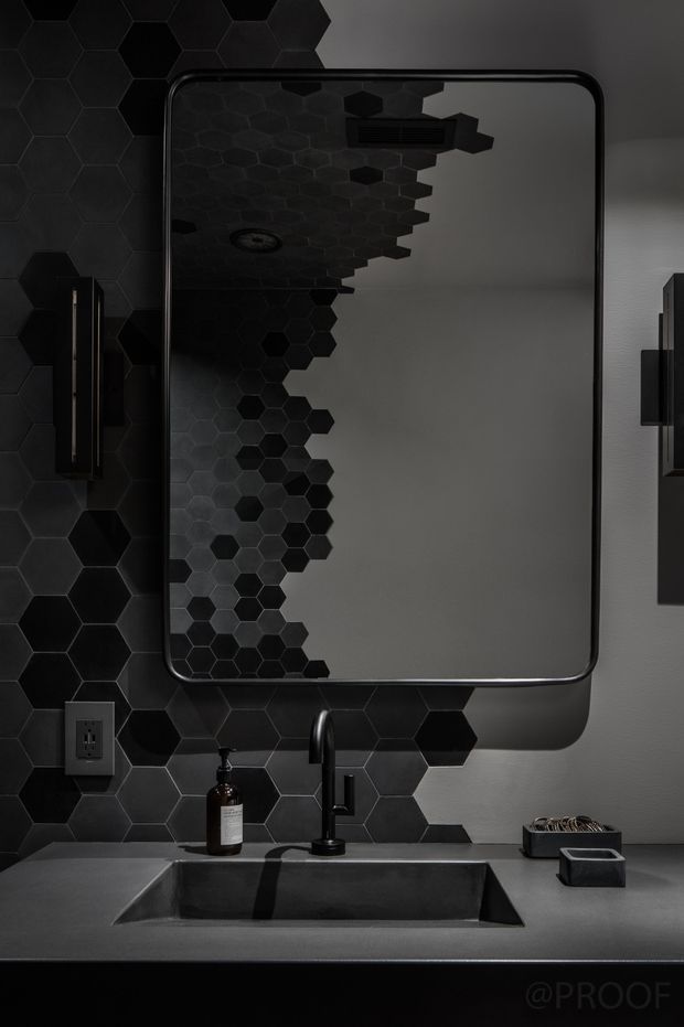 A moody contemporary bathroom vanity featuring a large rounded-rectangle mirror with a dark metal frame, a black hexagonal tile splashback, dark wall-mounted lighting, a dark rectangular basin, and a matte black faucet set into a grey benchtop. The mirror reflects the patterned tile wall and ceiling light, while minimal toiletries and accessories sit on the counter.