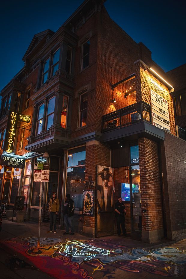 Night-time street view of a historic brick commercial building housing Ophelia's Electric Soapbox. The facade combines ornate Victorian-era bay windows and decorative trim with a newer brick-clad corner addition, illuminated signage, balcony, large windows, and a lively painted sidewalk and storefront frontage.