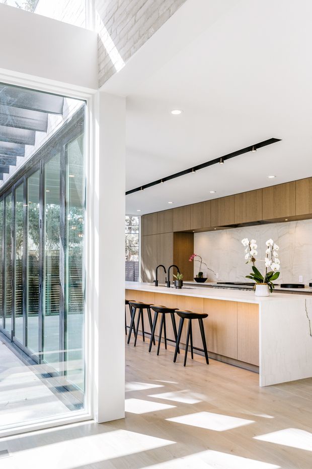 Bright contemporary kitchen viewed from an adjoining glazed corridor or sunlit space. The kitchen features a long island with a pale stone waterfall benchtop, integrated timber cabinetry, a pale marble-look splashback, black tapware, four black stools, and linear black track lighting. Full-height glazing and a high angled ceiling bring in strong natural light, with a glimpse of white-painted brickwork above.
