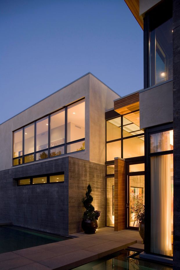 Twilight exterior view of a contemporary multi-level residence arranged around a narrow reflecting pool or water feature. The architecture features strong rectilinear volumes, dark masonry or concrete walls, smooth rendered upper walls, extensive black-framed glazing, and warm timber soffits and entry cladding. Interior lighting glows through the tall windows and glazed entry, creating a dramatic courtyard-like composition.