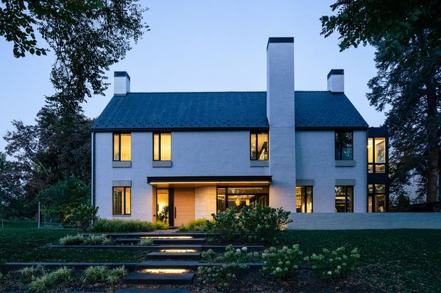 A two-storey modern residence photographed at dusk, with a symmetrical pale rendered facade, steep dark blue-black pitched roof, tall central chimney, smaller roof chimneys, and warm light glowing through the windows and recessed ground-floor entry. Broad landscaped steps lead up through a formal garden toward the house, framed by mature trees.