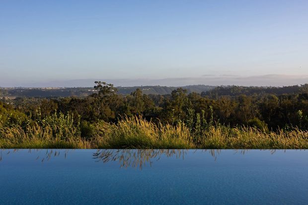 A tranquil landscape view across dense greenery and rolling wooded hills, reflected in the still surface of a dark blue infinity pool in the foreground. The image is dominated by natural scenery, with no visible building or interior.