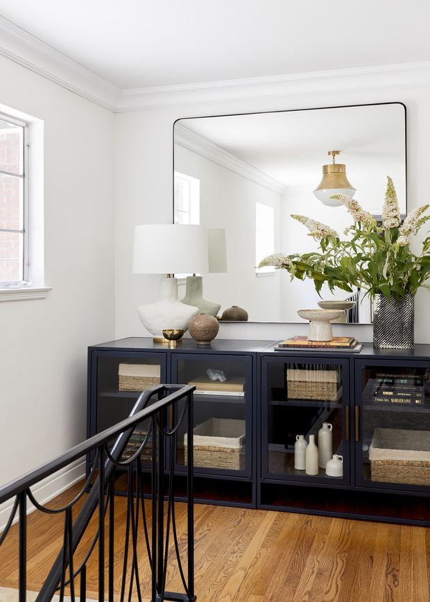 A refined residential landing or hallway vignette with a dark navy built-in glass-fronted console cabinet, warm timber flooring, white walls, ornate crown moulding, a large rounded-corner mirror, and carefully styled ceramic objects, books, a lamp, and a tall floral arrangement. A black metal stair railing occupies the foreground, indicating the space is adjacent to a staircase. The overall composition is bright, polished, and transitional in character.