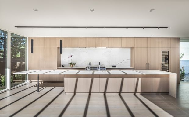 Contemporary open-plan kitchen with a large central island, extensive light timber cabinetry, a white marble-look splashback, integrated appliances, and floor-to-ceiling glazing. Strong linear shadows from the glazing fall across the pale floor, with glimpses of trees and water visible outside.