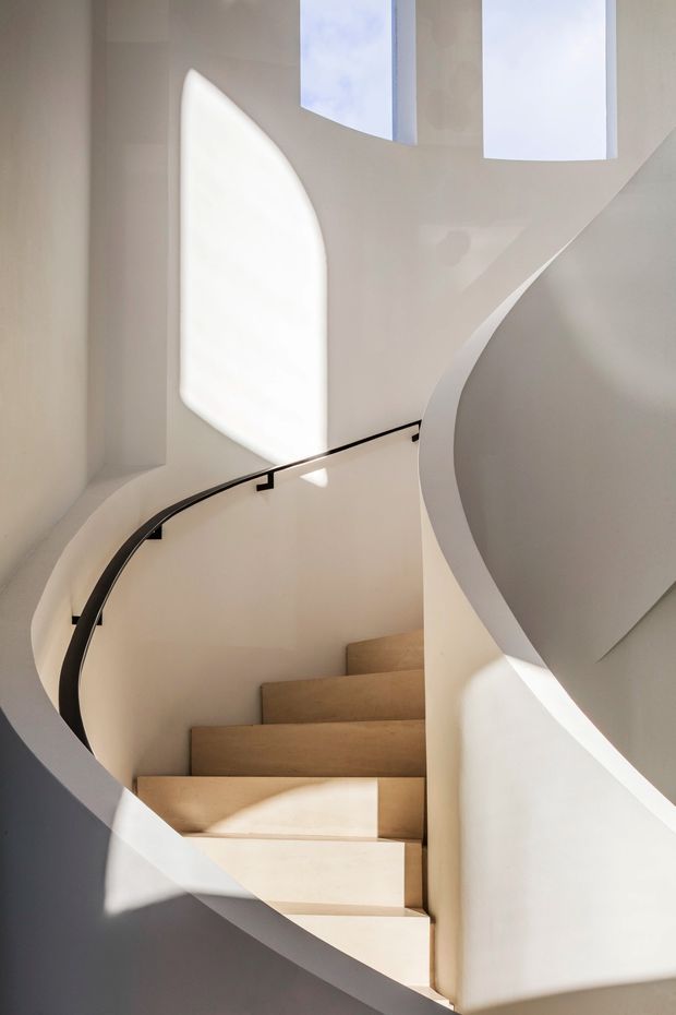 A sculptural curved staircase viewed from below, featuring pale timber or wood-finished treads, smooth white plastered curving walls and balustrade edges, and a slim dark handrail. Tall narrow openings above bring in strong natural light, creating dramatic geometric shadows across the minimalist stairwell.