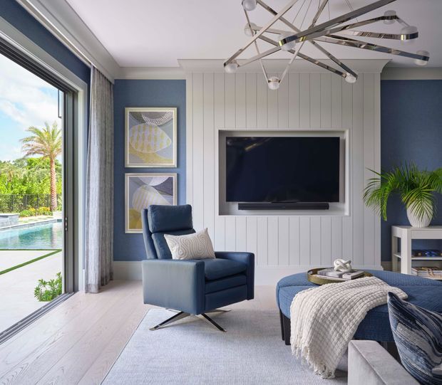 Bright, refined living room with a deep navy accent wall, white vertical wall panelling and a recessed wall-mounted television. The room features a blue upholstered armchair, matching ottoman and sofa seating, pale timber flooring, a large contemporary sculptural pendant light, framed artwork, curtains and a potted palm. Full-height glazing opens visually toward a swimming pool and garden outside.