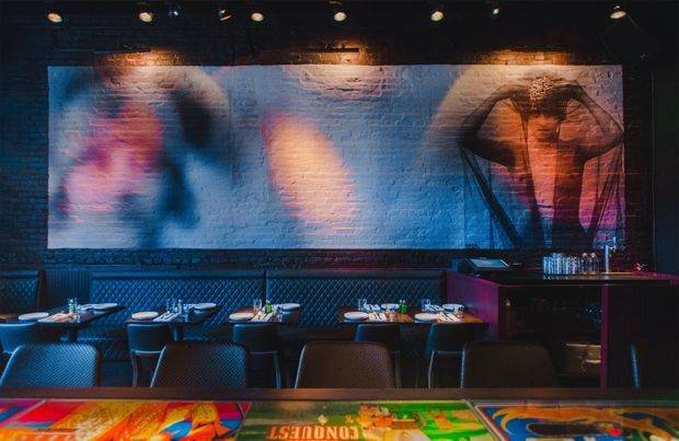 A dimly lit, atmospheric bar or restaurant interior with rows of dark upholstered booths and tables, a service counter at the rear, exposed brick walls, and large colourful projected or painted artwork. Warm amber and saturated blue, pink, and red lighting create a dramatic nightlife ambience. A colourful game or bar counter surface occupies the foreground.