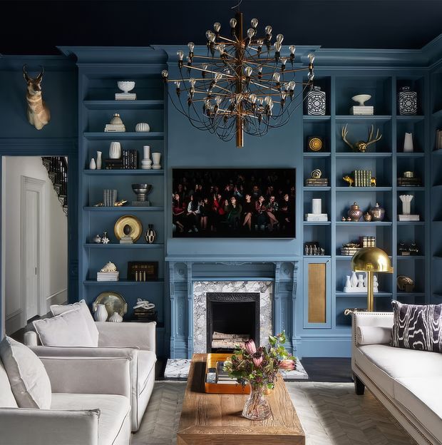 A richly styled living room with deep blue built-in shelving and cabinetry surrounding a television and ornate fireplace. Pale upholstered sofas frame a substantial timber coffee table, while a large sculptural chandelier hangs centrally. Decorative objects, books, artwork, and a mounted animal head fill the shelving, creating a refined, dramatic interior.
