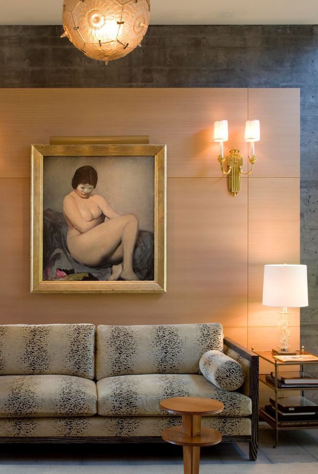 A richly styled living room vignette featuring a patterned upholstered sofa, a small tiered timber coffee table, layered table lighting, a decorative brass wall sconce, and a framed figurative artwork. The room combines warm timber wall panelling with a dark exposed concrete wall and polished decorative lighting, creating an eclectic, refined atmosphere.
