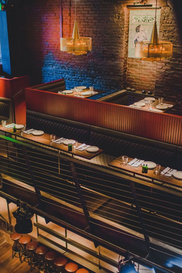 A moody, multi-level restaurant interior viewed from above, featuring rows of dark upholstered banquette seating and timber dining tables set with plates and glassware. Warm pendant lights hang against an exposed dark brick wall illuminated with blue, purple, and amber accent lighting. Metal railings, bar stools, and a vintage-style wall poster contribute to the urban hospitality setting.