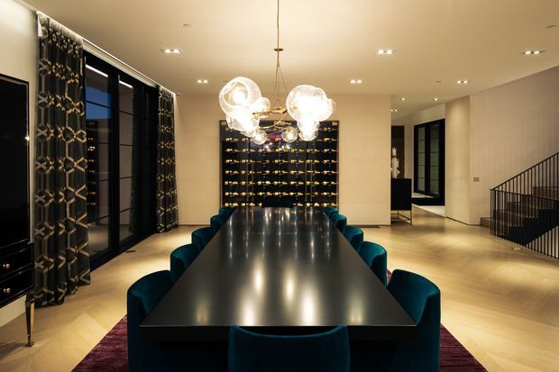 A sophisticated contemporary dining room with a long glossy black dining table surrounded by teal upholstered chairs. A sculptural cluster pendant light hangs above the table, while patterned dark curtains frame tall black-framed windows on the left. The room features pale timber flooring, warm neutral walls, recessed ceiling lights, a dark feature display wall at the rear, and a black metal stair balustrade to the right.
