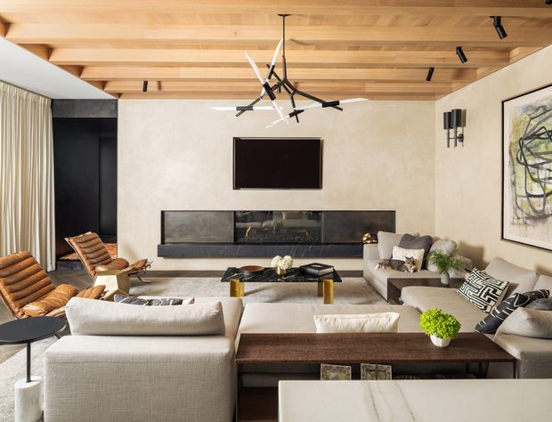 A refined contemporary living room centered on a large sectional sofa and low coffee table, with a wall-mounted television above a long dark built-in fireplace or media joinery unit. The room features a warm timber-lined ceiling with exposed beam-like detailing, textured light walls, dark built-in cabinetry, full-height curtains, sculptural black pendant lighting, and a large abstract artwork. Neutral upholstered furniture is accented by timber and black finishes.