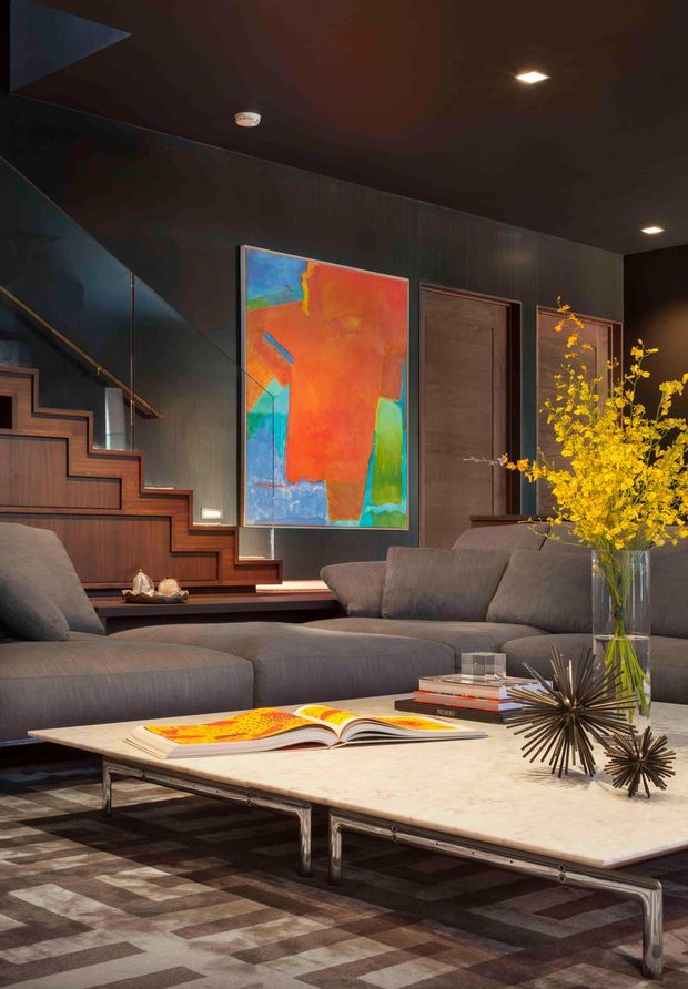 A dark, sophisticated living room with charcoal walls and ceiling, a large grey sectional sofa, and a low rectangular stone coffee table on a geometric patterned rug. A timber-lined staircase with open risers and a slim metal handrail rises along the left wall. The room features a vivid abstract artwork, timber door, recessed ceiling lights, sculptural dark starburst decor, books, and a tall vase of yellow branches. The overall composition is moody, refined, and contemporary with strong warm timber and colourful artistic accents.
