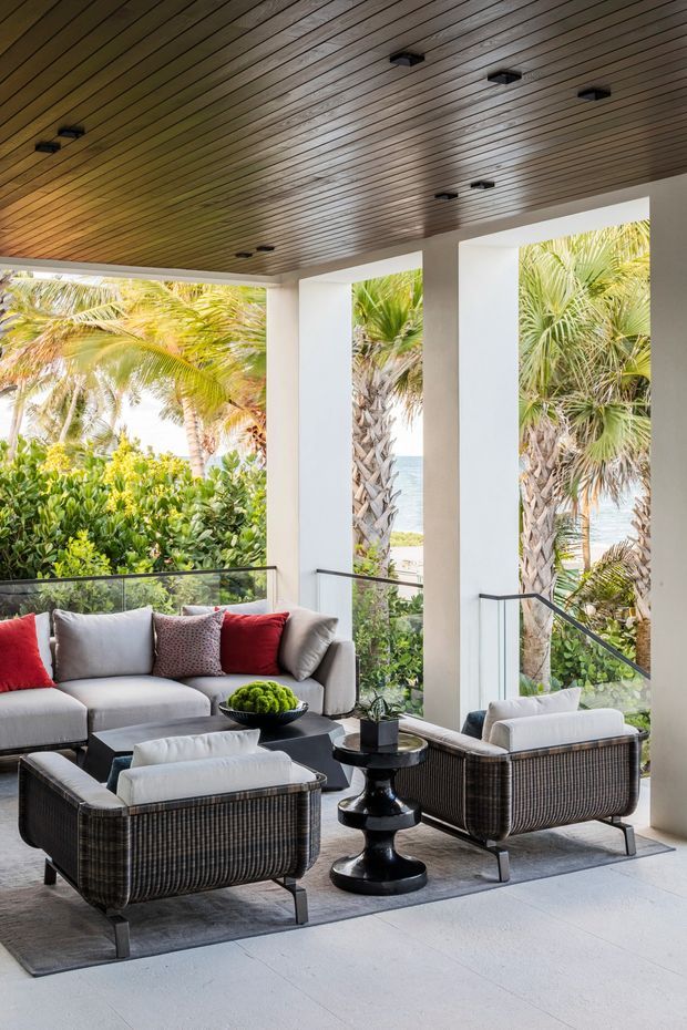 Bright, tropical-inspired living room with a large grey sectional sofa, two woven dark armchairs, a black pedestal side table, and a low black coffee table arranged on a grey rug. Tall open glazed bays framed by white structural columns overlook lush palms and a glimpse of the sea. The room has a warm slatted timber ceiling with recessed black spotlights and a pale large-format floor.