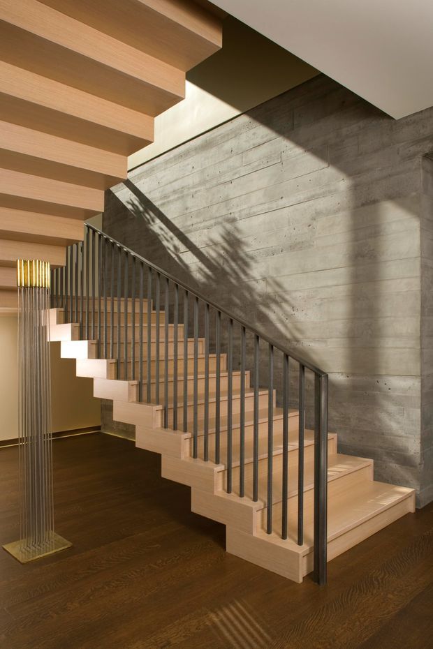 Interior view of a contemporary stairway with pale timber floating steps and matching overhead ceiling battens, set against a board-formed concrete wall. Slim dark metal balustrades, dark timber flooring, and strong sunlight create graphic shadows across the stair and wall. A sculptural floor lamp is visible at the lower left.