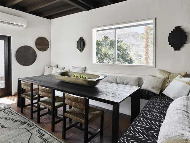 A bright, rustic-modern dining and living area with a long dark timber dining table, woven bar-height chairs, and a built-in upholstered bench layered with neutral cushions. White textured walls feature circular and geometric dark wall art, while exposed dark ceiling beams and terracotta-toned flooring add warmth. A large window overlooks trees and rocky hills, and a black-and-white patterned rug defines the seating area.