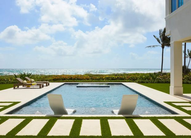 A contemporary beachfront pool terrace viewed from a manicured lawn. A rectangular swimming pool with submerged loungers occupies the foreground, bordered by pale geometric paving and clipped grass. Lounge chairs sit to the left, while part of a white multi-storey building with a covered columned terrace is visible on the right. Beyond a landscaped hedge is a bright ocean horizon with palm trees and scattered clouds.