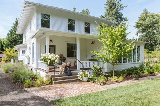 A bright exterior view of a two-storey white clapboard house with a covered front porch, decorative diamond-paned windows, white railings, and a landscaped garden. A brick-paved path and porch steps lead to the entrance, with mature trees and planting surrounding the property.
