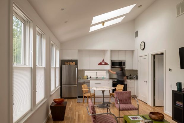 Bright, compact open-plan kitchen and dining/living area with tall windows along one wall, white built-in cabinetry, stainless-steel refrigerator, timber flooring, skylights, and a small dining table with mid-century-inspired seating. A red pendant light and patterned lounge chairs provide accents.