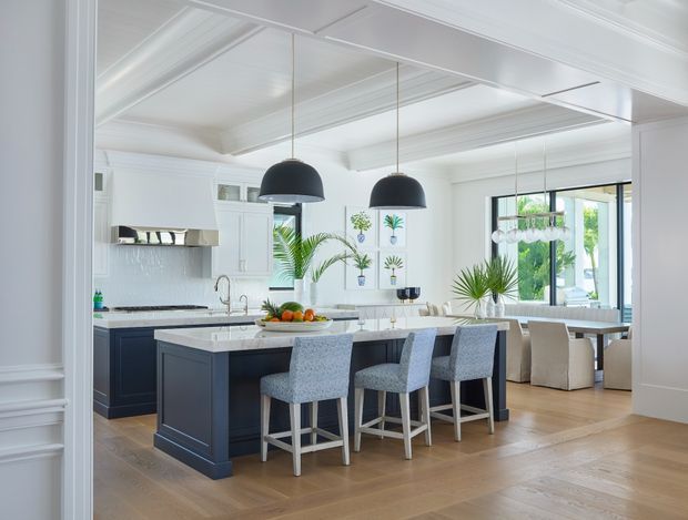 Bright, spacious open-plan kitchen and dining area with a large dark island, pale stone benchtop, four upholstered bar stools, white cabinetry, and black dome pendant lights. The room features white coffered or panelled ceilings, light timber flooring, a tiled splashback, framed botanical artwork, and abundant natural light. A dining table and chairs are visible beyond the island.