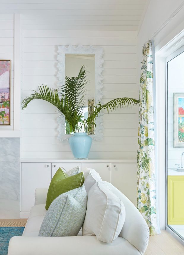 Bright coastal-style interior vignette featuring a white shiplap-paneled wall, ornate white-framed mirror, palm plant in a pale blue vase, white sofa with patterned green and blue cushions, and floral curtains beside a glazed door or window. White built-in cabinetry and a glimpse of a pale green kitchen area are visible to the right.