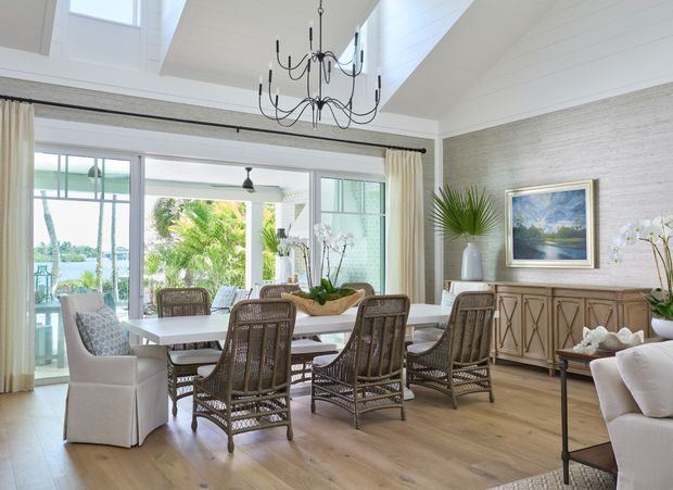 Bright coastal-style dining room with a large timber dining table surrounded by woven rattan chairs and upholstered end chairs. The room features light timber flooring, textured pale wall panelling, white shiplap ceiling, expansive glazed doors opening to a covered outdoor area, a dramatic black multi-arm chandelier, and a timber sideboard with decorative detailing. Tropical planting and water are visible beyond the glazing.