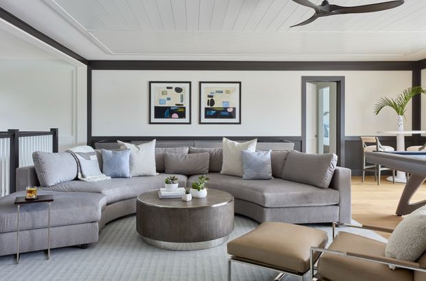A bright, refined living room with a large curved grey sectional sofa, layered neutral cushions, a round dark coffee table, and a soft area rug. The room features dark grey wall trim, framed artwork, white painted plank ceiling, expansive glazing, and a contemporary ceiling fan. A dining area is visible through the opening at right.