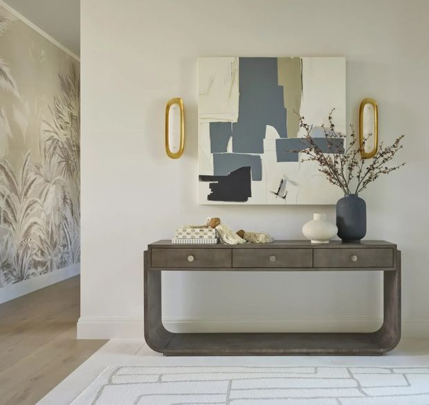 A refined entryway or hallway vignette featuring a dark wood console table with rounded ends, a large abstract artwork, sculptural gold wall sconces, a ceramic vase, and decorative branches. A patterned botanical wallpapered area and pale timber flooring are visible to the left, with a light geometric rug in the foreground.