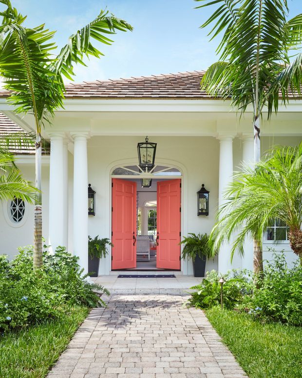 Bright exterior view of a tropical, columned entrance facade. A paved walkway leads through lush palm landscaping to an open pair of coral-red front doors beneath a deep portico, with white stucco walls, decorative columns, lantern sconces, an arched transom window, and a tiled pitched roof.