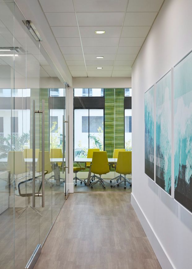 Bright contemporary office corridor leading into a glass-enclosed meeting room. The space has full-height glass partitions and doors with metal handles, a suspended white tile ceiling with recessed lighting, light wood-look flooring, white walls, large windows, and vivid yellow-green office chairs around a meeting table. Abstract turquoise artwork lines the right wall.