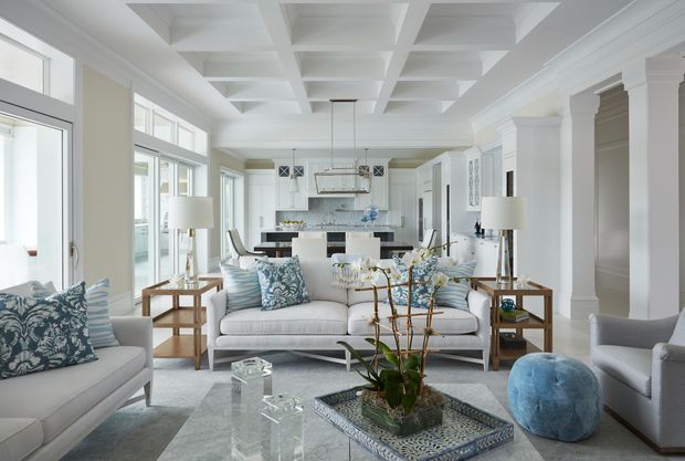 Bright, refined open-plan living room with a large coffered ceiling, extensive windows, white classical trim and columns, and a white kitchen visible beyond the seating area. The room features pale upholstered sofas and armchairs, blue patterned cushions, glass coffee tables, a patterned area rug, and a central dining or kitchen island with pendant lighting.