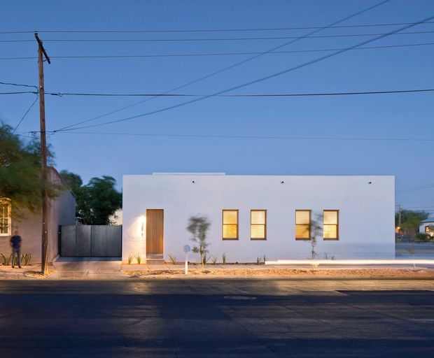 A simple single-storey, flat-roofed white building viewed from across a street at dusk. The rectangular facade has a recessed timber-toned entry door, several warm-lit rectangular windows, minimal detailing, and a narrow landscaped strip along the sidewalk. Utility poles and overhead wires are visible in the surrounding urban streetscape.