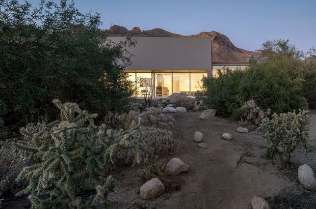 Exterior view of a low-profile contemporary house at dusk, partially screened by dense desert planting and boulders. The building has a broad flat-roofed form, a simple dark wall volume, and extensive warm-lit glazing facing a rugged rocky mountain landscape.