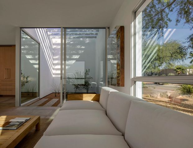 A bright, minimalist living room with a large cream sectional sofa, extensive floor-to-ceiling glazing, and views into a planted courtyard. Sliding glass doors open to a narrow glazed outdoor courtyard or sunroom with patterned sunlight, timber flooring, and built-in timber cabinetry. The space features white walls and ceilings, warm timber floors and joinery, and strong indoor-outdoor visual connections.