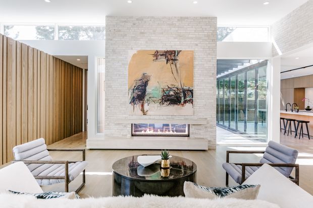 Bright contemporary living room with a white-painted brick fireplace wall, linear built-in gas fireplace, abstract artwork, large clerestory windows, and extensive glazing opening toward a glazed outdoor or courtyard area. The room features a round dark coffee table, light upholstered lounge chairs, a pale sofa, timber flooring, and a vertical timber batten feature wall. A kitchen with island seating is visible beyond the glazed opening.