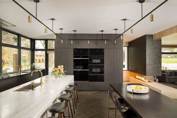 A sophisticated contemporary open-plan kitchen and dining space with a large white stone island, integrated dark cabinetry and appliances, black-framed floor-to-ceiling windows, brass pendant lights and tapware, and a dark timber dining table. A linear fireplace is built into a dark feature wall beside warm timber seating. The room is bright, spacious and refined, with views to landscaped grounds through the glazing.