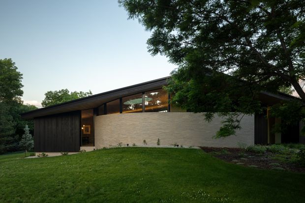 A low-slung contemporary residential house set within a landscaped garden, viewed from the front exterior. The building features a broad single-pitched roof, dark vertical timber cladding, expansive clerestory-style glazing, and a curved pale brick or masonry wall forming the main entry facade. Mature trees frame the house and a lawn occupies the foreground.
