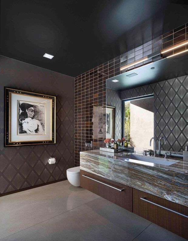A dramatic contemporary bathroom featuring a long double vanity with a richly veined stone benchtop, dark wood cabinetry, twin basins and brushed metal tapware. Dark charcoal walls are covered with diamond-patterned textured wallpaper or panels, while a vertical section of small square tiles frames the large mirror. A wall-hung toilet, framed artwork, recessed ceiling lights and linear LED lighting complete the moody, luxurious interior.