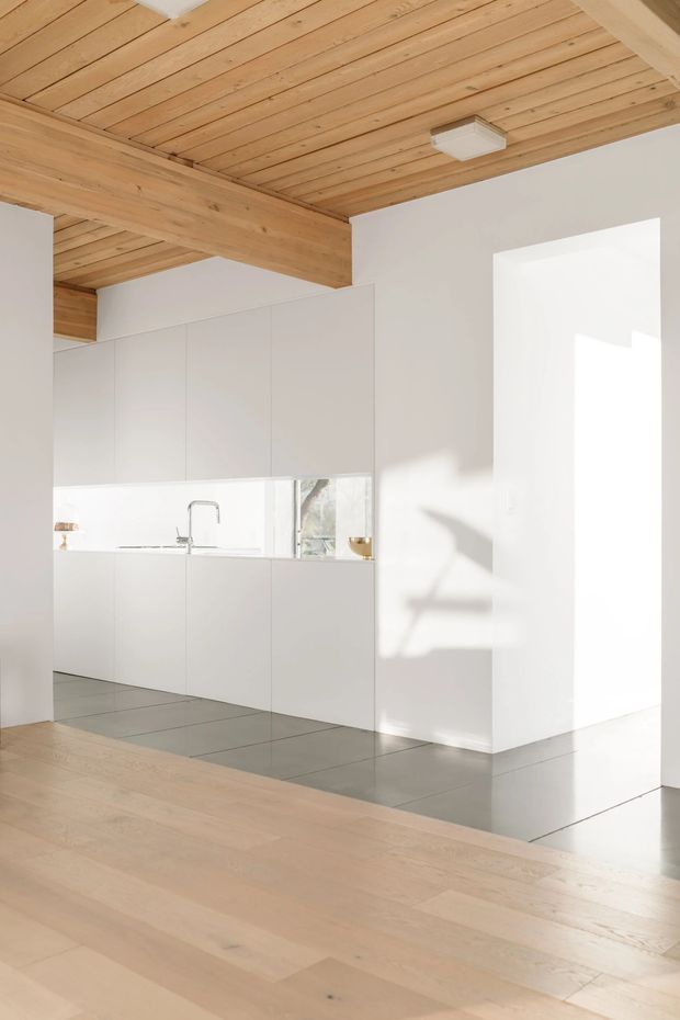 Bright minimalist open-plan kitchen viewed from an adjoining living area. The kitchen features handleless white full-height cabinetry, a slim reflective backsplash, integrated sink and tap, and a pale grey tiled floor. The adjoining area has light timber flooring, while the room is defined by a warm exposed timber plank ceiling and substantial timber beams. Strong natural light enters from the right, creating crisp shadows on the white walls.