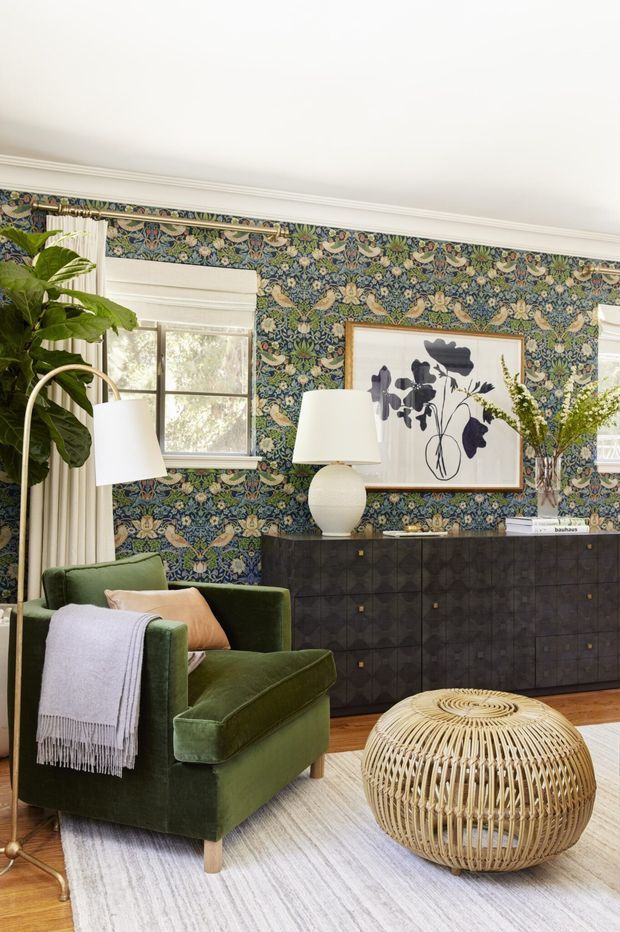 A richly styled living room with dark botanical patterned wallpaper, white decorative ceiling moulding, wood flooring, and a large pale rug. The room features a deep green upholstered armchair with a draped throw, a woven round ottoman, a dark built-in sideboard, botanical artwork, lamps, curtains, and indoor plants. The overall aesthetic is eclectic and traditional with strong vintage-inspired pattern and natural textures.