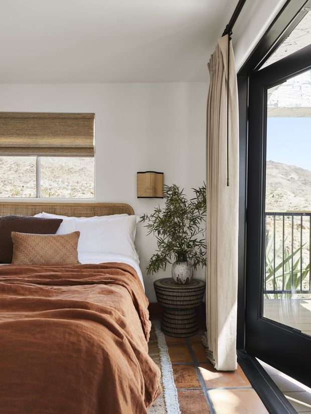 A warm, compact bedroom with a cane/rattan bedhead, white bedding, layered rust and terracotta blankets, and a dark accent cushion. A small woven wall light and potted plant sit beside a textured dark pedestal table. The room has white walls, terracotta-toned tiled flooring, a woven roller blind, full-height black-framed glazed doors, and neutral linen curtains. Bright desert-like terrain is visible beyond the glazing and balcony railing.